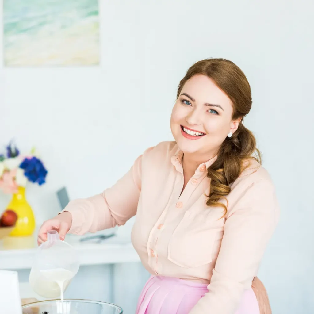 Smiling female pastry chef pouring milk into a bowl in a bright kitchen.
